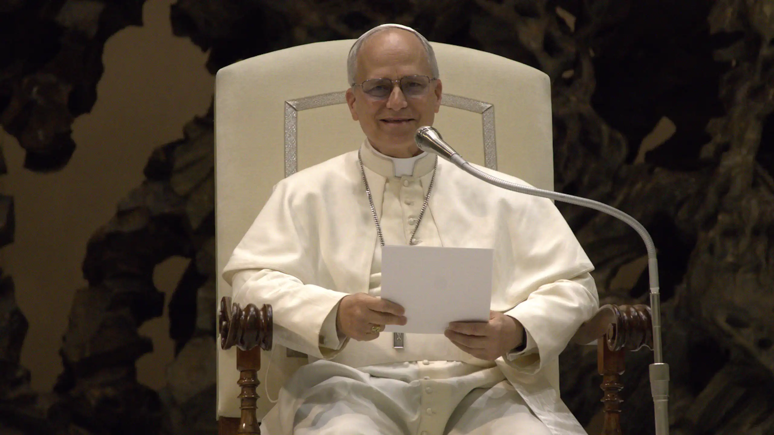 A seated man dressed in white robes holding a piece of white paper and speaking into a microphone