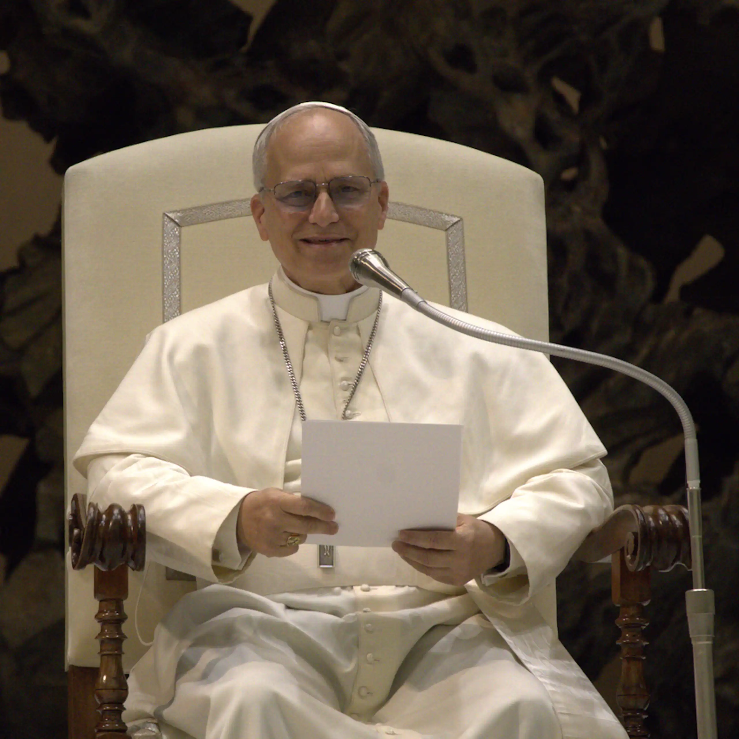 A seated man dressed in white robes holding a piece of white paper and speaking into a microphone
