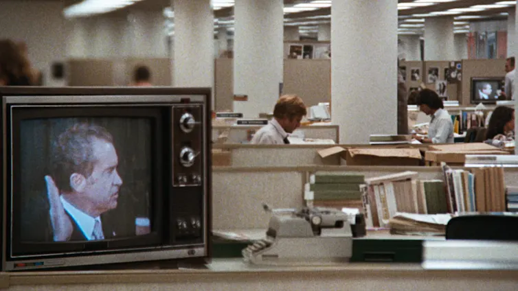 Una televisión muestra a Richard Nixon en medio de una redacción periodística mientras dos hombres trabajan al fondo.