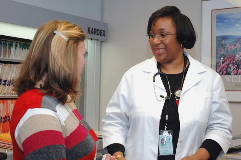 A doctor wearing a white medical coat talks with a patient in a striped sweater.