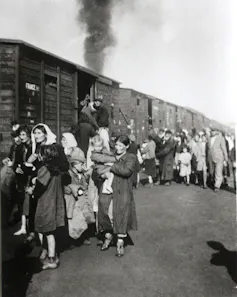 A black and white photo shows a crowd of people, many of them women and children, gathering outside rickety wooden train cars.