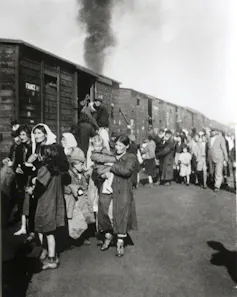 A black and white photo shows a crowd of people, many of them women and children, gathering outside rickety wooden train cars.