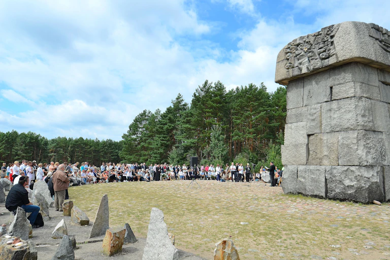 A large stone monument stands in grass, with people standing on the edge of the grass and woods in the distance.