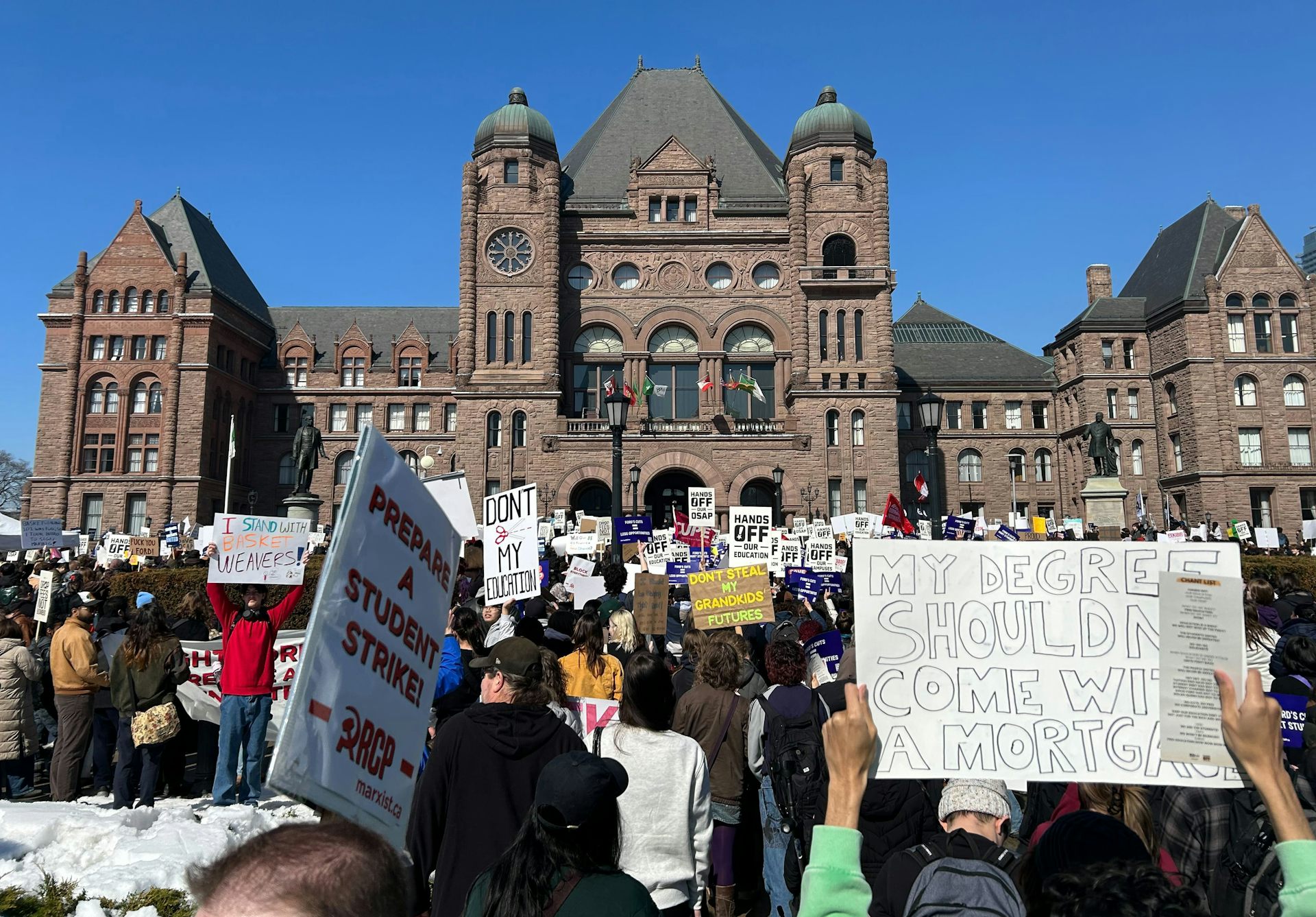 People stand with protest signs, one that says 'my degree shouldn't come with a mortgage' outside a large stone building.