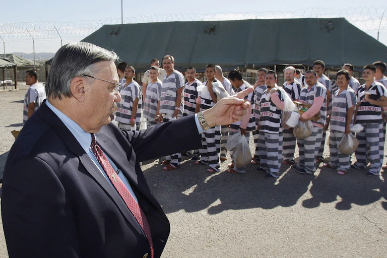 A man in a suit points to several prison inmates standing outdoors.