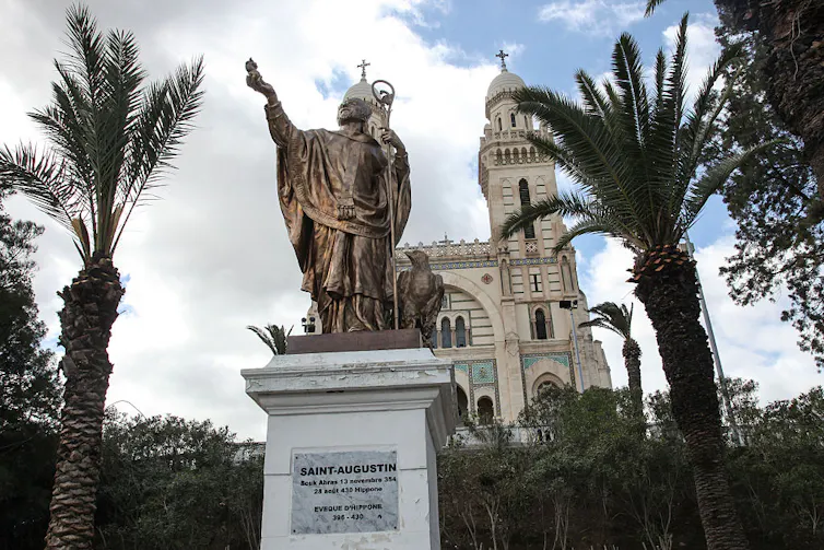 A large statue on a pedestal shows a man reaching up toward the sky, with palm trees and a tall church tower in the background.
