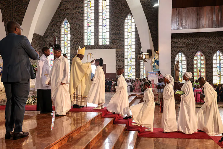 A line of people in white robes stand before a man in priestly robes and a white hat at the front of a church.
