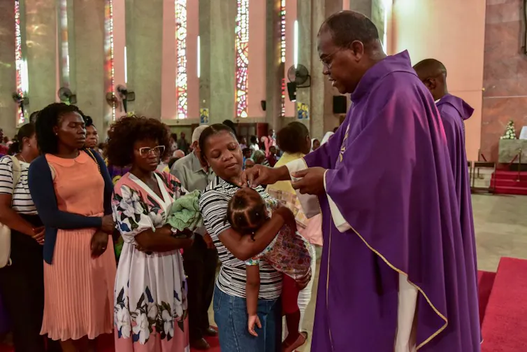 A man in a purple robe smudges ashes onto the forehead of a toddler held by a woman, with several other women waiting behind her.