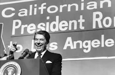 A man at a podium in a jacket and tie gestures to a crowd.