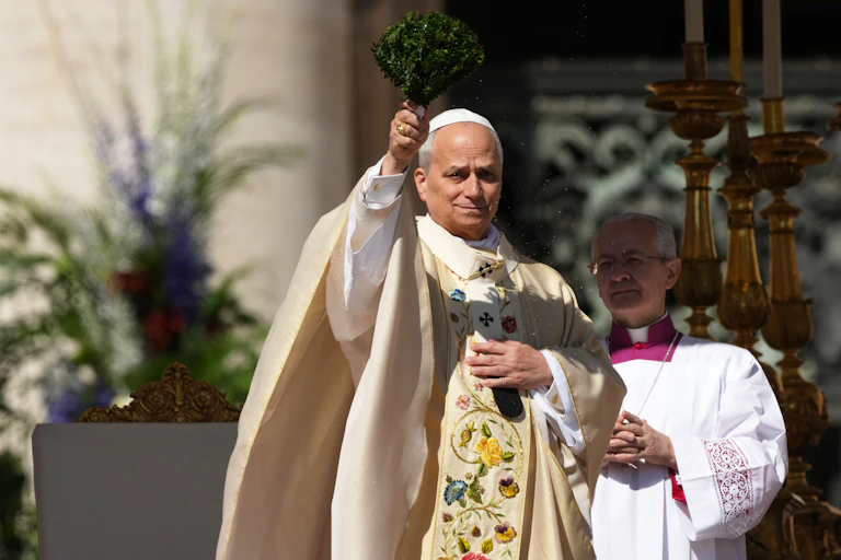 A man in white robes with embroidery holds up a bunch of green leaves as he stands outside.