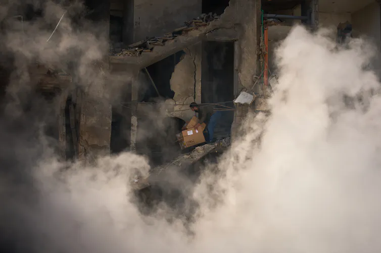 A man stands in a destroyed building as smoke rises around him.