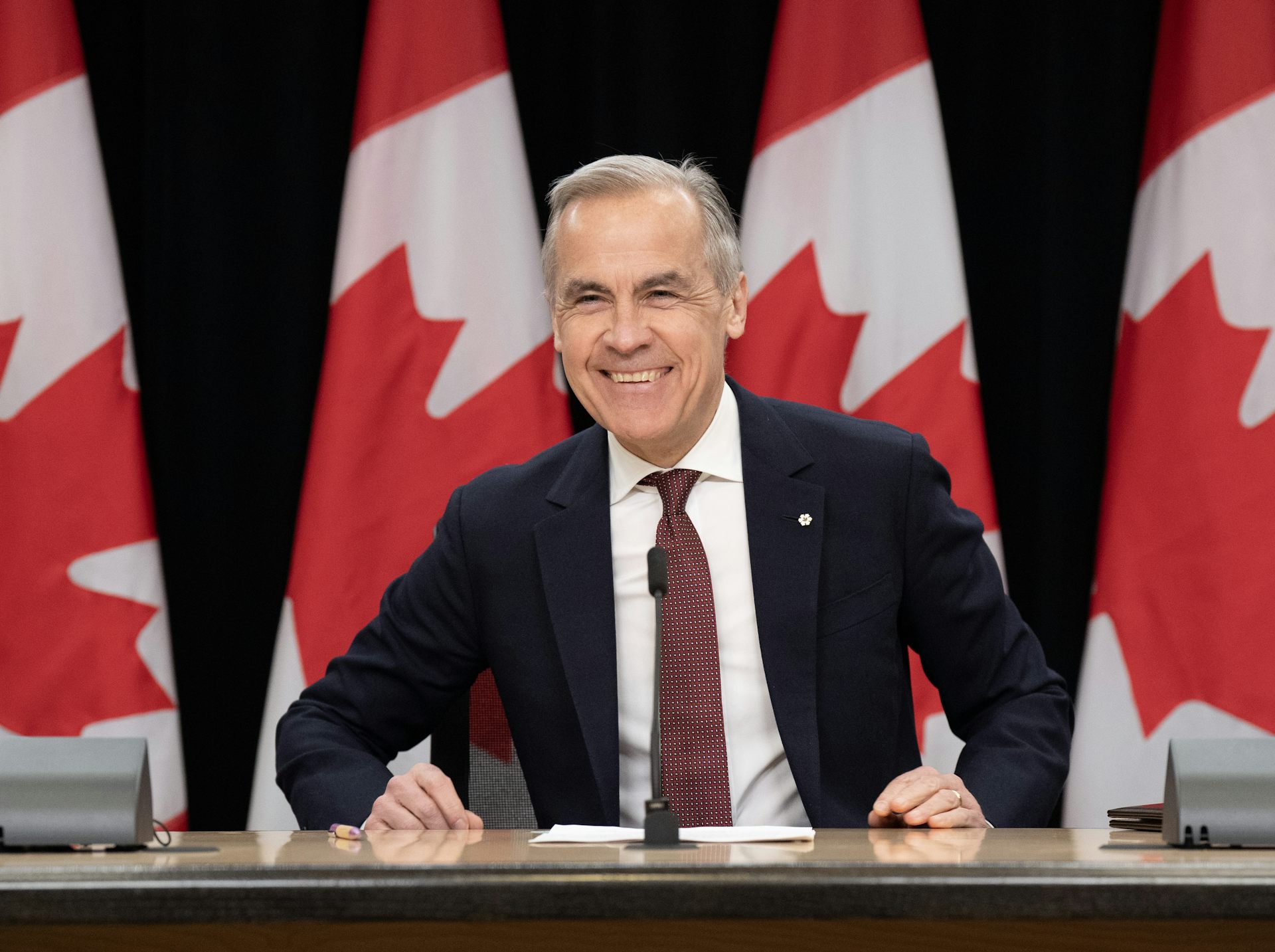 Un hombre con cabello corto y gris sonríe ampliamente mientras está sentado en una mesa con una hilera de banderas canadienses detrás de él.