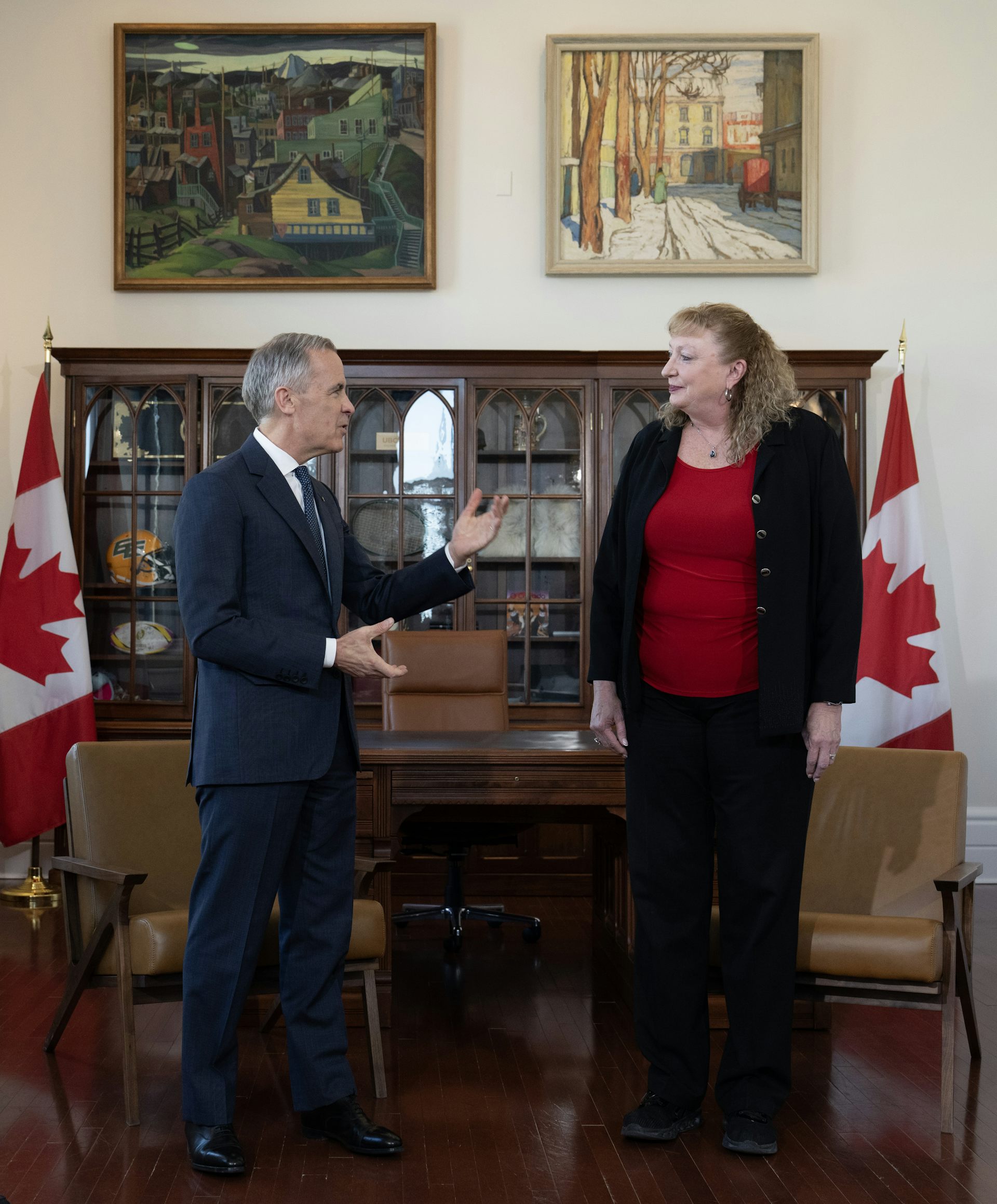 Un hombre delgado con cabello corto y gris está hablando con una mujer alta con cabello rubio oscuro y rizado en la oficina.