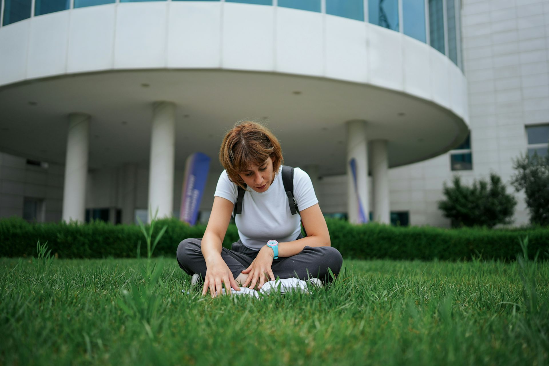 Une jeune femme lit ou ouvrage assise sur l'herbe devant une institution universitaire