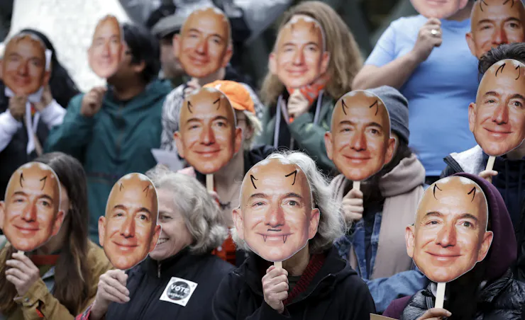 People hold small cardboard images of Amazon CEO Jeff Bezos in front of their faces.