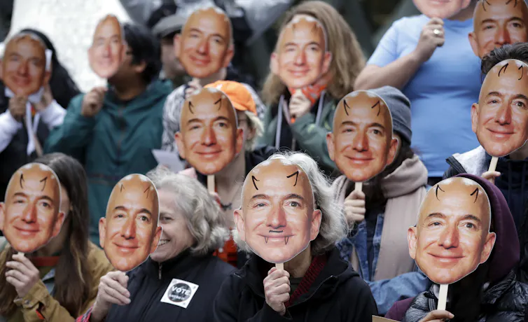 People hold small cardboard images of Amazon CEO Jeff Bezos in front of their faces.