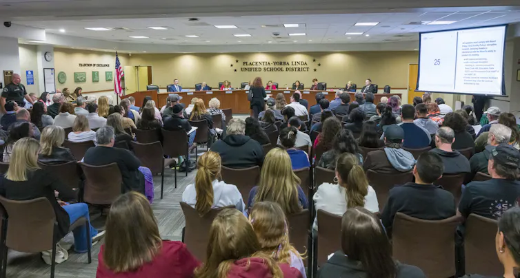 A large group of people are seen seated in a room with a projector, facing toward a row of people seated side by side at a table.