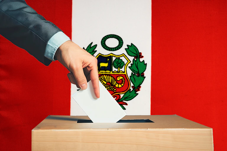 Una mano deposita una papeleta en una urna con la bandera de Perú al fondo