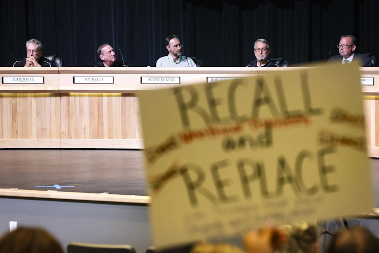 A hand-drawn sign that says 'Recall and Replace' is seen right below a group of five people seated at a large wood table.