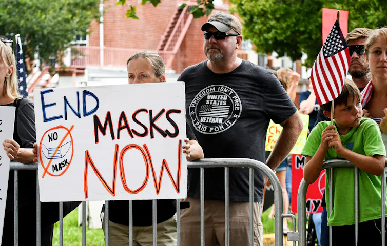 People stand along a metal barricade and one woman holds a sign that says 'End masks now.' A boy next to her holds a small American flag.