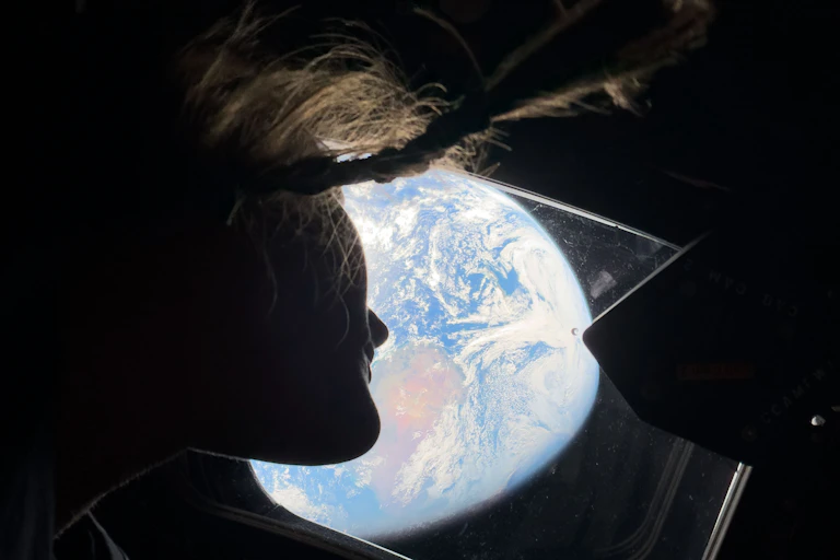 An astronaut looking out a window from below. The Earth is taking up most of the view outside the window.