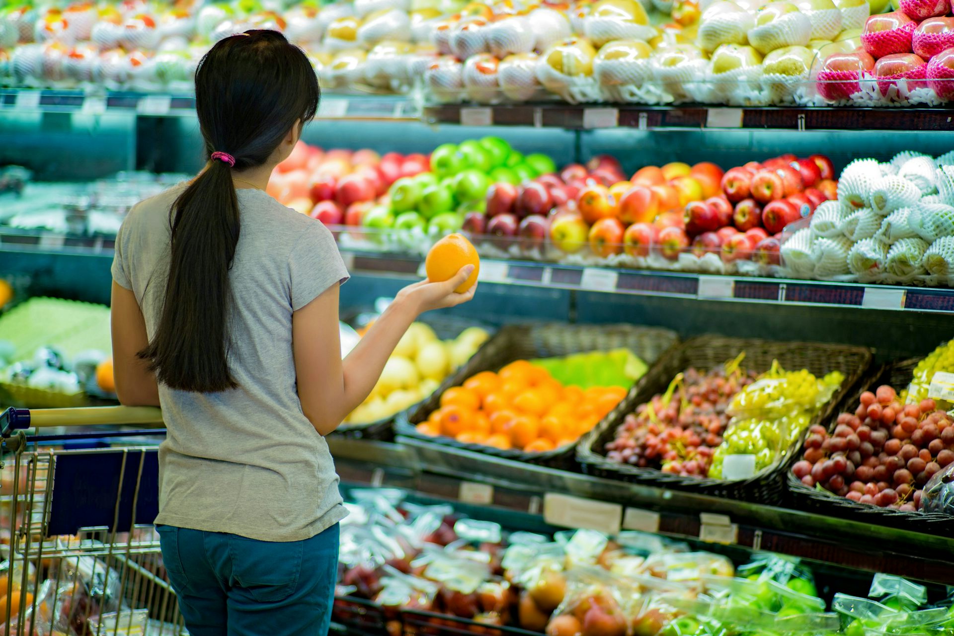 A person with their hair in a ponytail faces away from the camera toward shelves of fresh produce, holding an orange in their outstretched hand