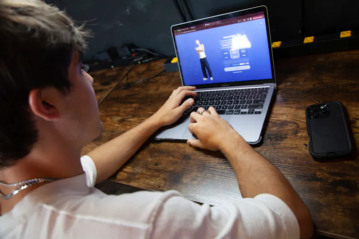 A young man seen from behind looks at a laptop screen with an image of a young man in a white shirt and black pants.