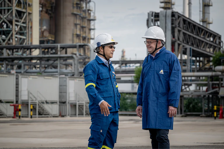 Two men in white hard hats and navy protective jackets at an oil refinery