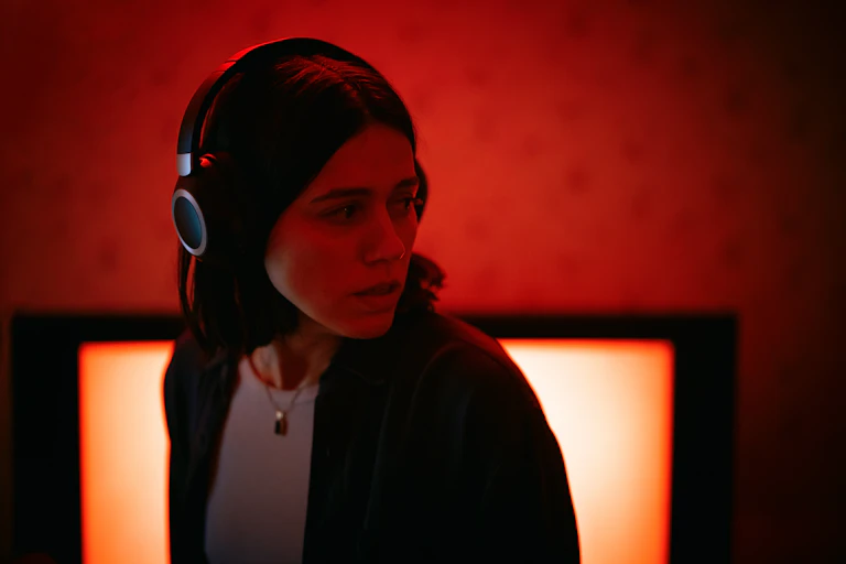A woman in headphones against a red wall looking to the right, unnerved.