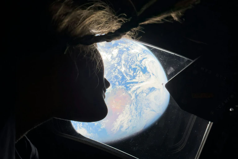 An astronaut looking out a window from below. The Earth is taking up most of the view outside the window.