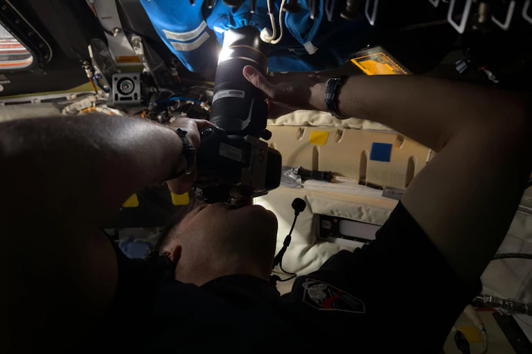 An astronaut inside a spacecraft holding a camera up to a window with a cloth covering. The lens is pushed through a hole in the covering.