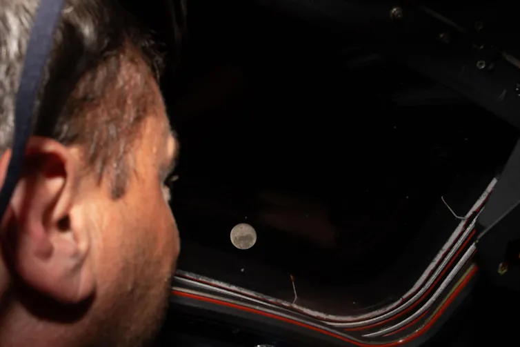 An astronaut looking out the window of the Orion spacecraft, where the full moon is visible in space.