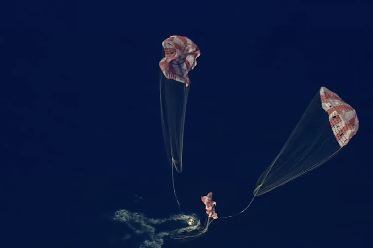 A small crew capsule floating in the ocean, with three parachutes attached to it falling down towards the water.