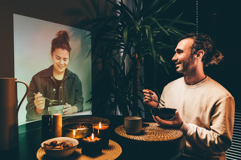 A brunette man in a sweatshirt holds a bowl as he smiles, looking toward a woman's image projected on a screen.
