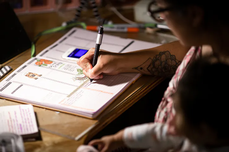 A woman holds a pen as she writes in an agenda book, sitting bent over a table.