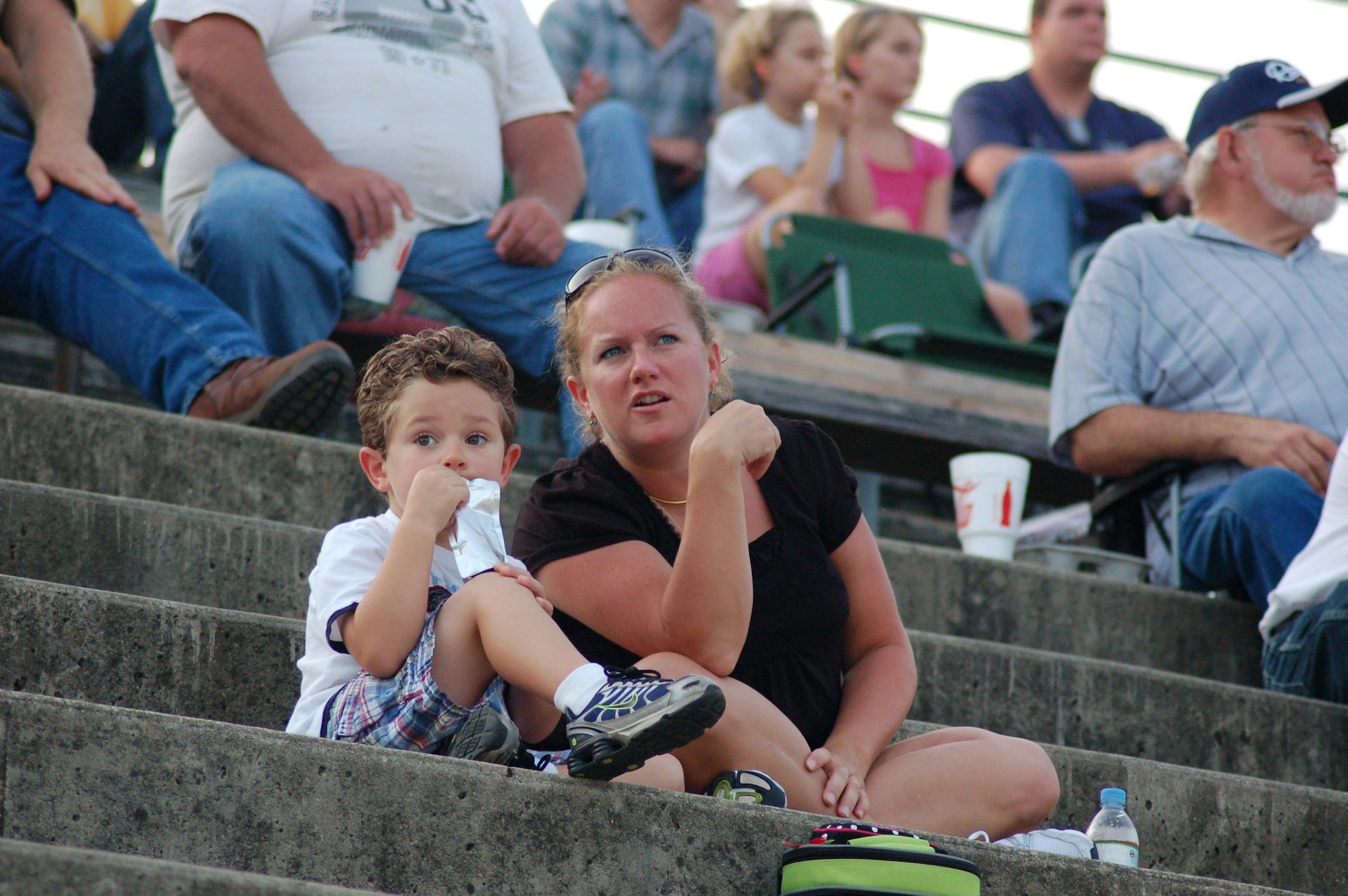 A woman with blonde hair talks to a young child while the two sit in the stands of a motor speedway.