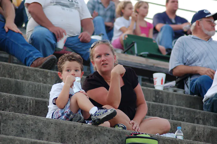 A woman with blonde hair talks to a young child while the two sit in the stands of a motor speedway.