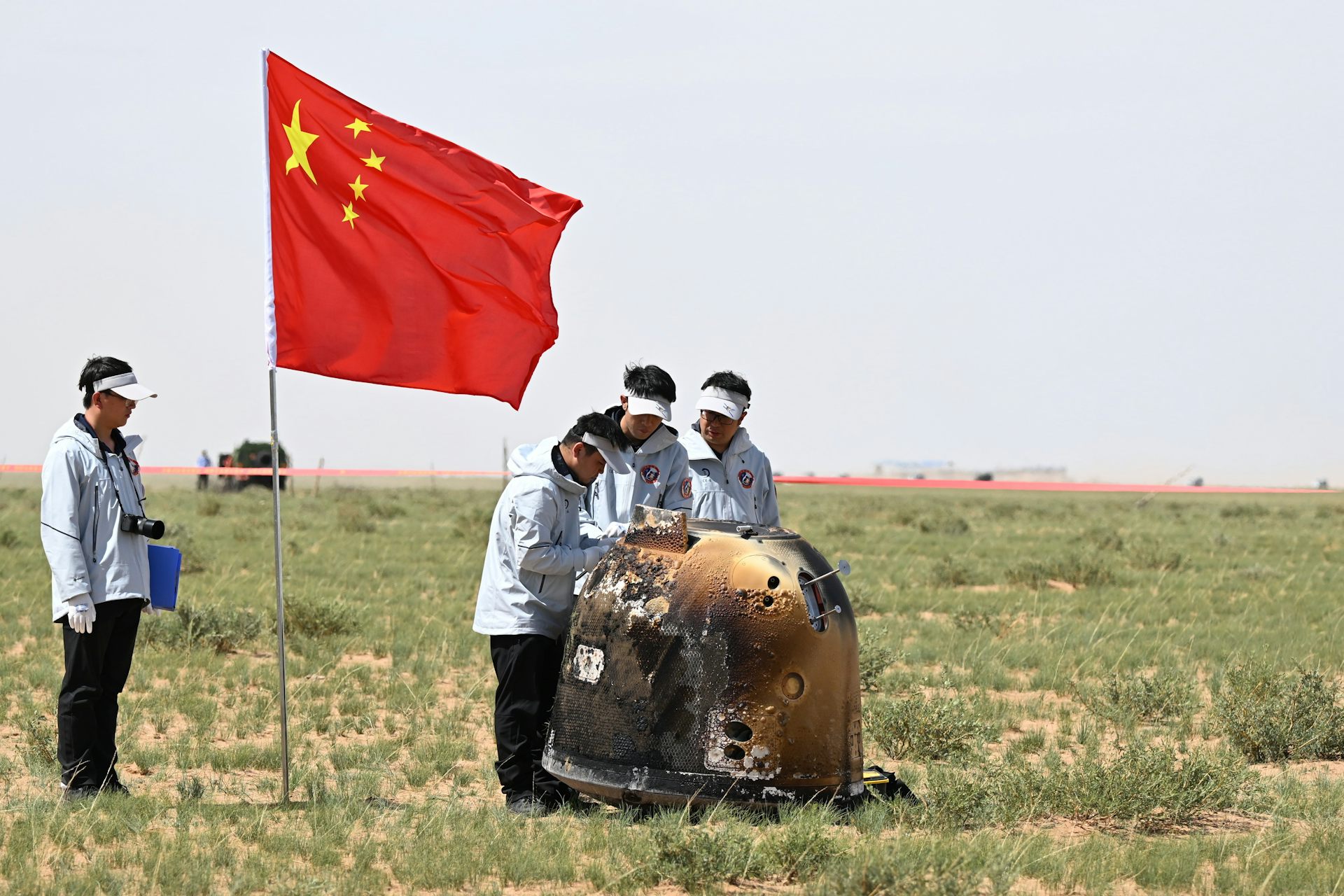 People gathered around a metal capsule in a field, flying a Chinese flag.