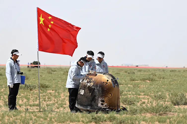 People gathered around a metal capsule in a field, flying a Chinese flag.