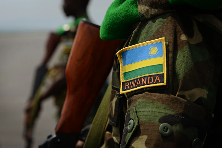 An arm in the foreground of a man dressed in military fatigues with the map of Rwanda on the sleeve, and holding a gun