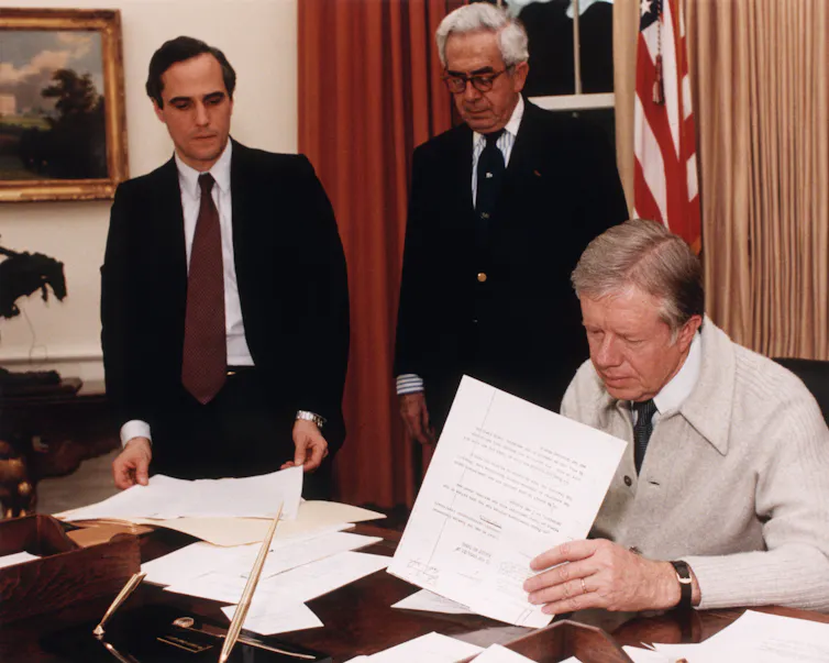 A man at a desk with two men standing behind him as he signs a piece of paper.