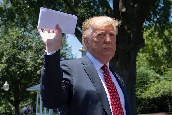 A man in white shirt, red tie, blue jacket holds up a folded piece of paper.