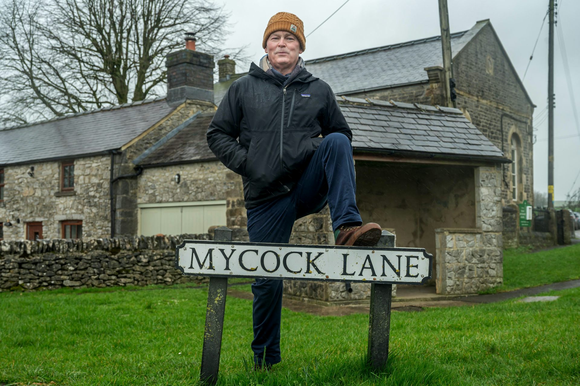 A man standing with one foot on a sign that reads 'Mycock Lane'.