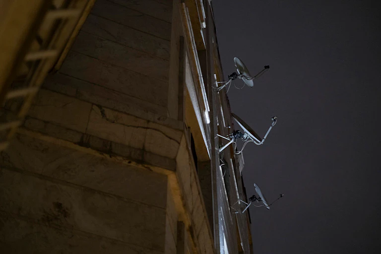 An apartment building with three satellite dishes hanging out of windows, illuminated against the night sky.