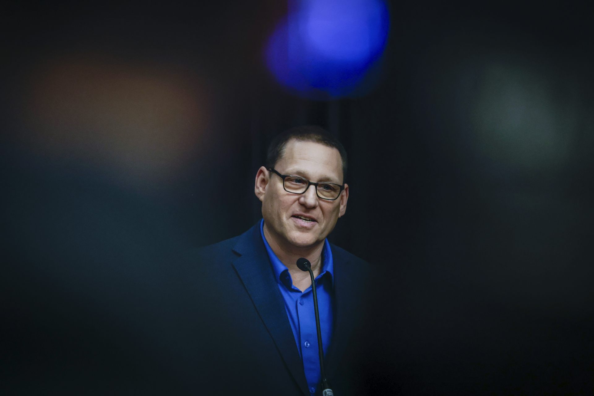 A man with short hair and glasses at a news conference; a blue light glows over his head.