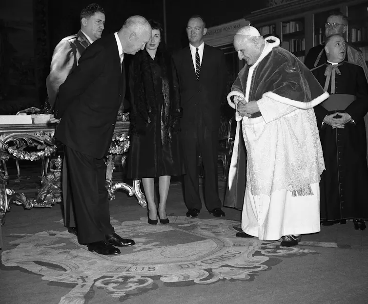 A man in a black suit and another in white priestly robes bow toward each other, as some others stand quietly in the background.