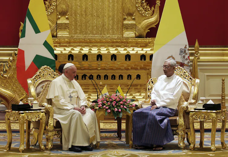 Two men, one in priestly robes, sit facing each other on ornate golden chairs, with flags behind them set against a gilded backdrop.