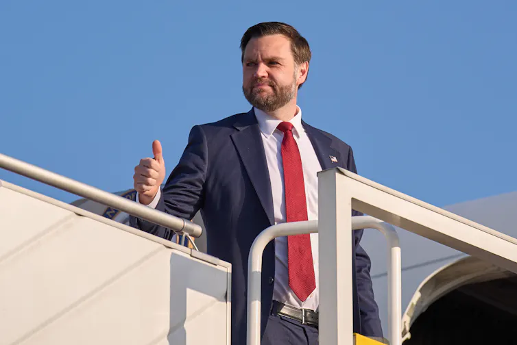 a man with dark hair and a beard gives a thumb's up as he boards an airplane