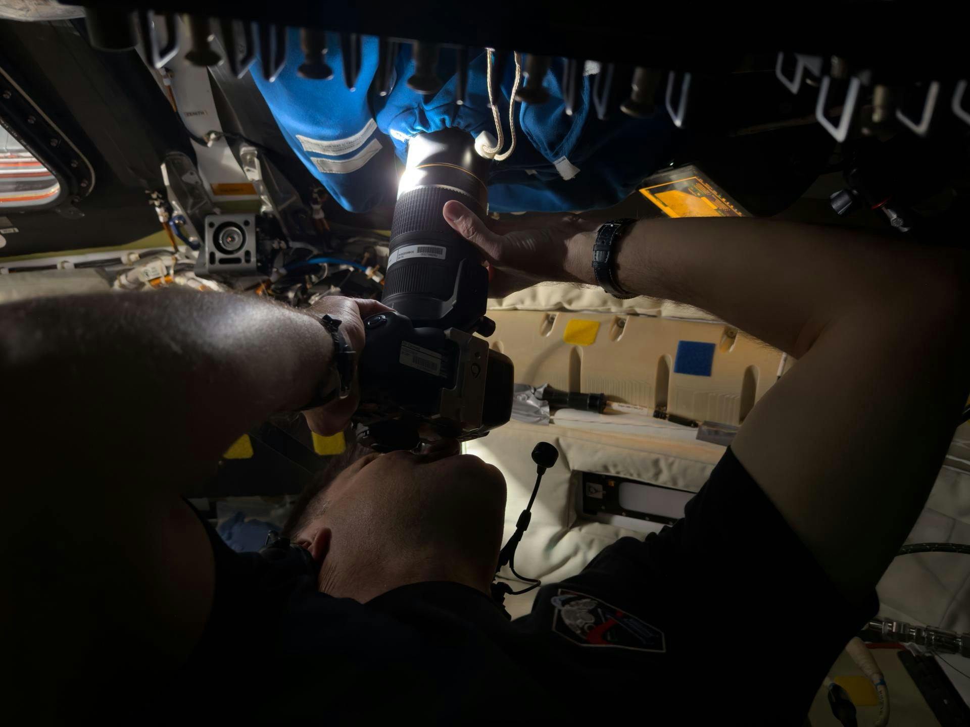 Um astronauta dentro de uma nave espacial, segurando uma câmera encostada a uma janela coberta por um pano. A lente é enfiada por um orifício na cobertura.