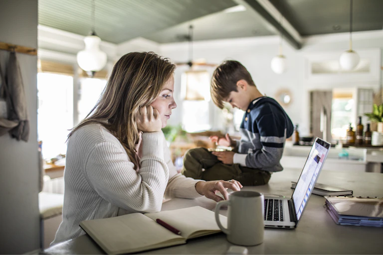 Woman works on laptop with her son sitting on the table next to her